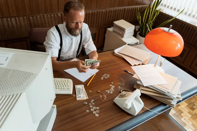 Man with beard in vintage office counting cash and coins at desk with retro computer and phone. A pilot plan for payroll research tools before any subscription