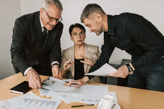 Three adults engaged in a dynamic discussion at an office table, analyzing documents and planning strategies. Budgeting for productivity software means pricing the end at the beginning