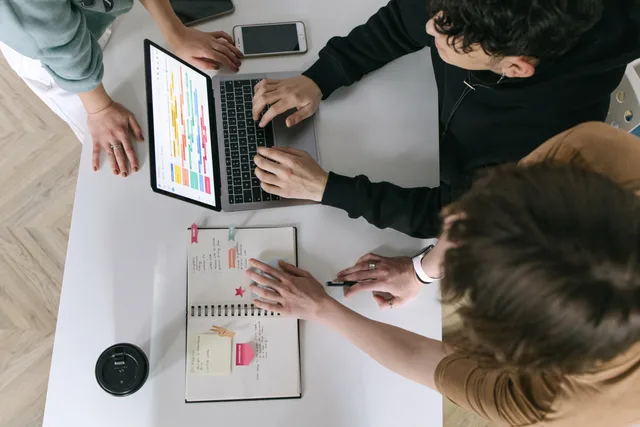 A high-angle view of a team collaborating over a laptop and notebook in a modern office. Business productivity software comparison: Planner, Asana and Trello on one shared workflow, with permissions, exceptions and export